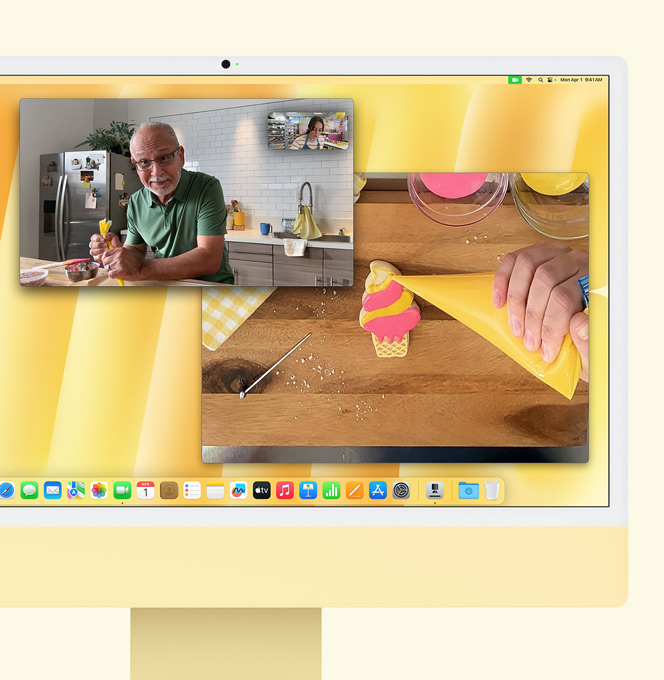 iMac jaune. Démonstration de Desk View. L’écran montre une pâtissière participant à un appel vidéo avec une autre personne. Une fenêtre montre la pâtissière et le visage de l’homme. L’autre fenêtre montre les mains de la pâtissière en train de faire un glaçage.