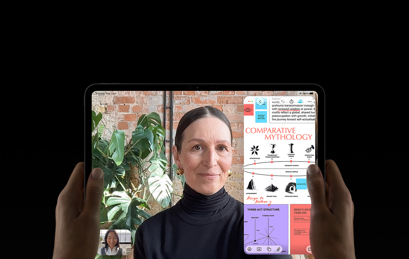 iPad Pro, front exterior, space black color, left and right hands holding in landscape orientation, rounded corners, black display bezel, display showing a FaceTime conversation being live translated, and an educational app showing a presentation of 'Comparative Mythology' with text, diagrams and handwritten notes
