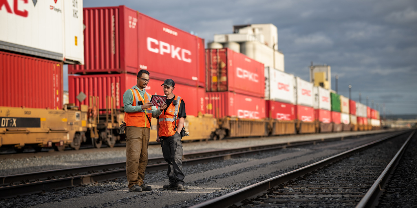 Zwei Bahnarbeiter blicken auf ein iPad, während sie auf einem Bahnhof neben einem stehenden Zug stehen, der Schiffscontainer transportiert