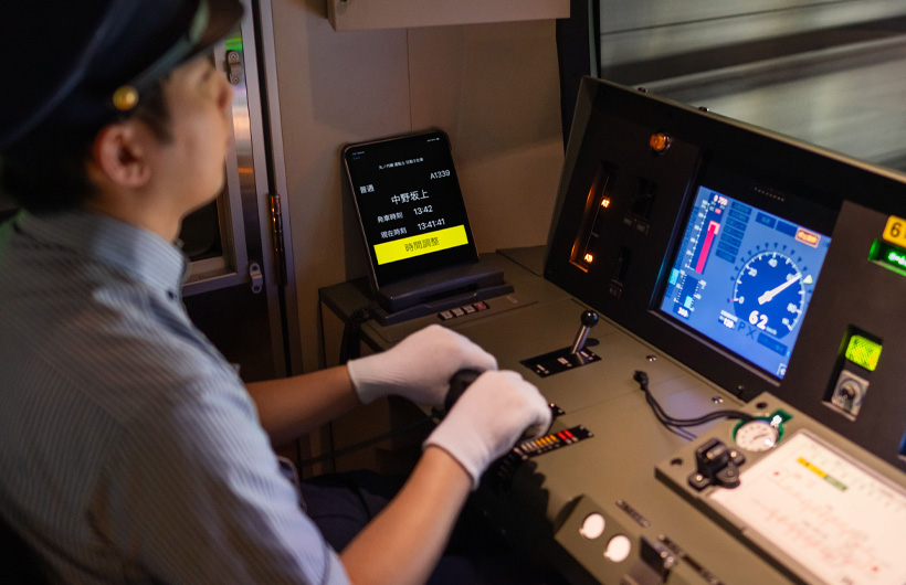 An iPad displaying stations and times rests near the control panel, where a train operator works.