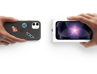 En hånd holder en iPhone 11 med silikone-etui med klistermærker på bagsiden, ved siden af holder en anden hånd en æske med en ny iPhone 17e