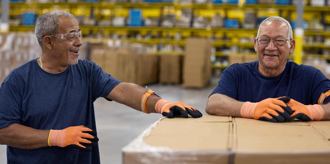 Employees wearing safety glasses and gloves stand by a pallet of boxes in a warehouse