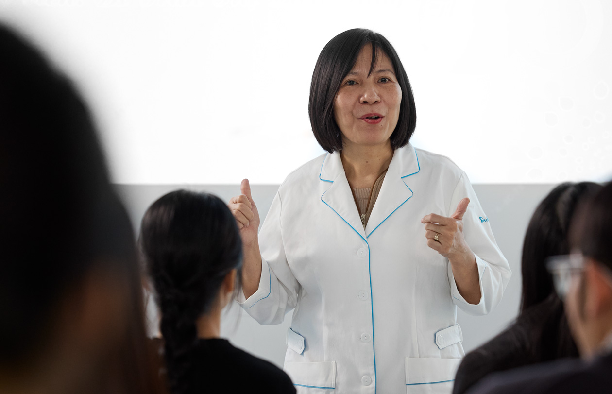 An instructor in a white lab coat teaches at the front of a classroom full of students