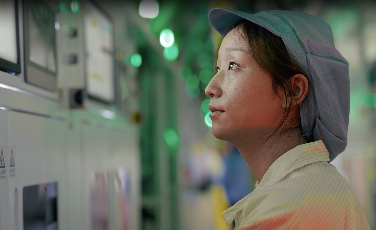 A uniformed employee works in a green-lit component manufacturing facility