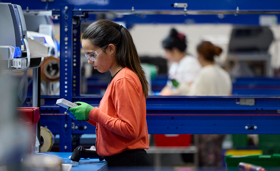 An employee wearing safety gloves and glasses inspects a production component at an upright work station